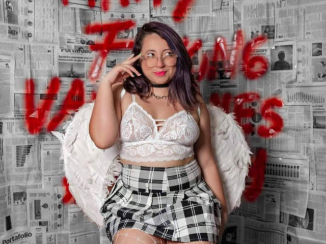 CristinaSmit with purple hair, glasses, white lace bra, plaid skirt, and feather wings, touching her face, smiling, wearing a black choker, showing her tits.