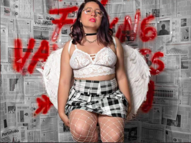 CristinaSmit has purple hair, glasses, white lace bra showing her boobs, plaid skirt, fishnet stockings, white wings, black choker, and red lipstick, standing confidently.