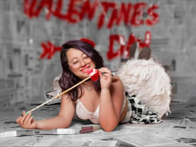 CristinaSmit with purple hair, smiling, eating a red heart on a stick, wearing white angel wings and a plaid skirt, lying on newspaper, her tits in a white lace top.