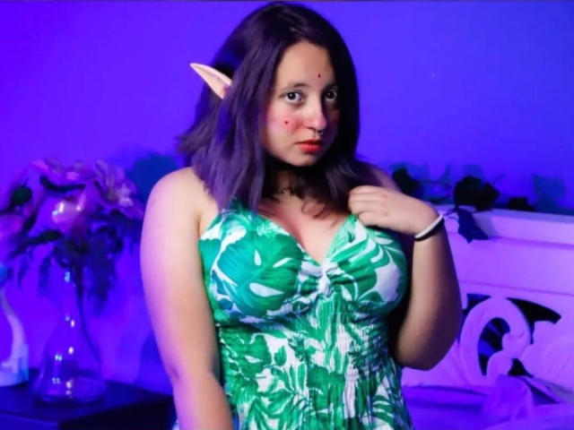 CristinaSmit with elf ears, wearing a green floral dress, has big tits, purple-pink face, and dark wavy hair, standing in a blue-lit room with flowers in background.