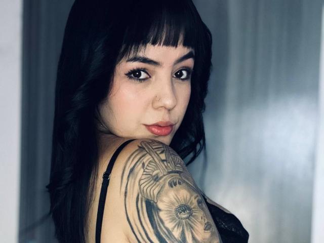 AlyssaVelascuria with black hair, nose ring, and tattoo on her shoulder, looking over her shoulder, showing her tits in a black lace top.