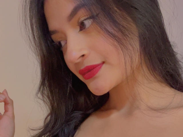 LizzyMarhall69 with long black hair, red lipstick, and a slight smile, looking to the side with her right hand raised. Her brown eyes and fair skin are visible.