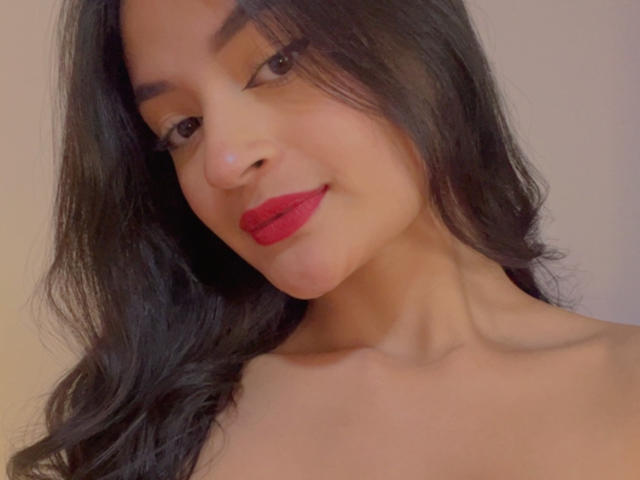 LizzyMarhall69 with long black hair, red lipstick, and brown eyes, smiling slightly, topless, showing her bare shoulders and neck, in a soft-lit selfie.