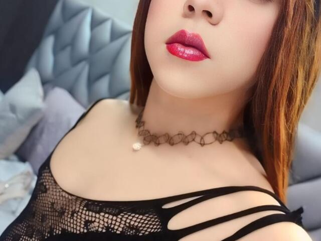 AndrewTaylor69 with shiny pink lips, fair skin, and red hair, wearing a black mesh top with strap details, and a delicate choker necklace. Boobs visible through the mesh.