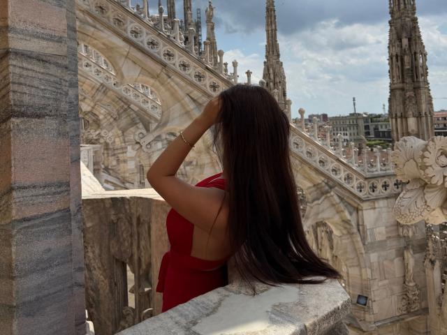 MollyMoo, con lunghi capelli castani e un vestito rosso, in piedi con una mano sulla testa, guarda una cattedrale gotica con guglie appuntite, indossando un braccialetto dorato, in una giornata di sole..