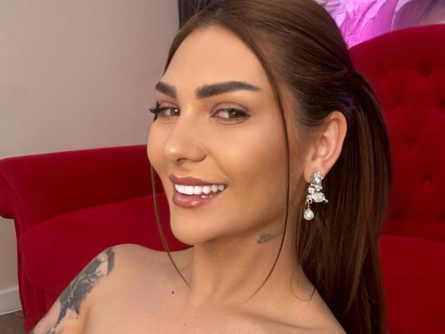 AnissaDesire, avec une queue de cheval brune, portant des boucles d'oreilles en diamant, souriant avec des lèvres roses brillantes, montrant ses seins avec un tatouage sur son épaule gauche. Canapé rouge en arrière-plan..