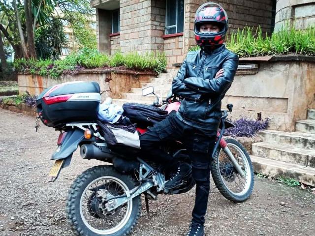 Egonga stands with arms crossed, wearing black leather jacket, black pants, and red helmet, next to a red motorcycle with a black top case, on a gravel path.