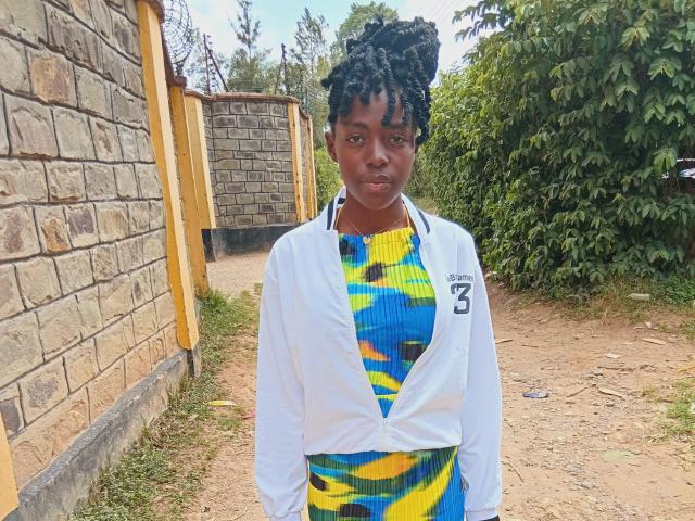 Shaznesh with dark skin, medium build, and curly black hair, wearing a white jacket over a colorful yellow, blue, and green patterned dress, standing on a dirt path.