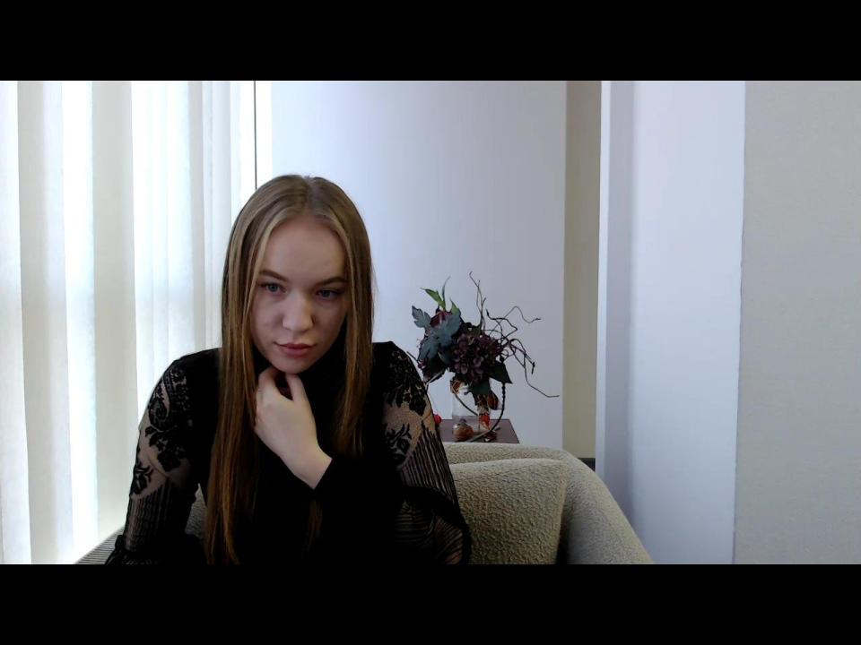 Violettia with long blonde hair, black lace top, hand on chin, looking slightly bored or sad, sitting on beige chair, small floral vase in background.