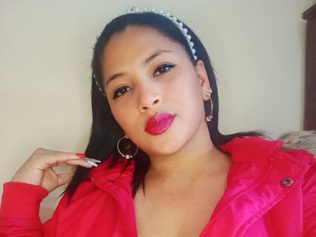 RoxaneMilf with dark hair, wearing a red jacket, hoop earrings, and a headband, has glossy pink lips and is touching her hair with manicured nails.