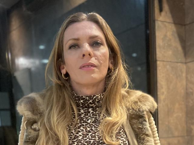 AnaliceColin, a middle-aged woman with long blonde hair, stands in a city at night. She wears a leopard print turtleneck and a fur-trimmed coat. Her face shows light makeup, and she has small gold earrings. Her expression is neutral, slightly pouty. The image focuses on her upper body, with a reflective glass surface in the background. The lighting is even, highlighting her facial features and coat texture. Her hair is slightly messy, and her skin is fair. The composition centers on her face.