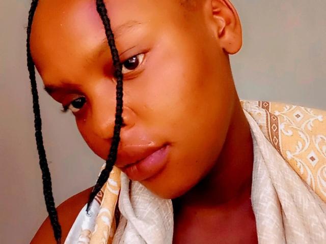 Tinna-hot with a bald head and black braided hair, brown skin, and a subtle smile, wearing a patterned shirt with a white towel draped over shoulders.