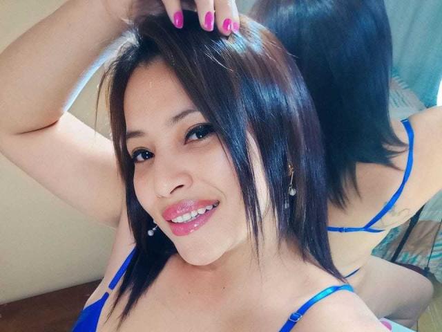 RoxaneMilf with black hair, pink lipstick, blue bra, and earrings, smiling at mirror, hand in hair, pink nails, reflection shows back, small tits, cute, sexy.