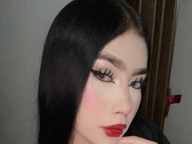 BellaDalthon has long black hair, heavy eyeliner, long lashes, pink blush, red lipstick, and a small nose piercing. She's looking to the side with a slightly pouty expression.