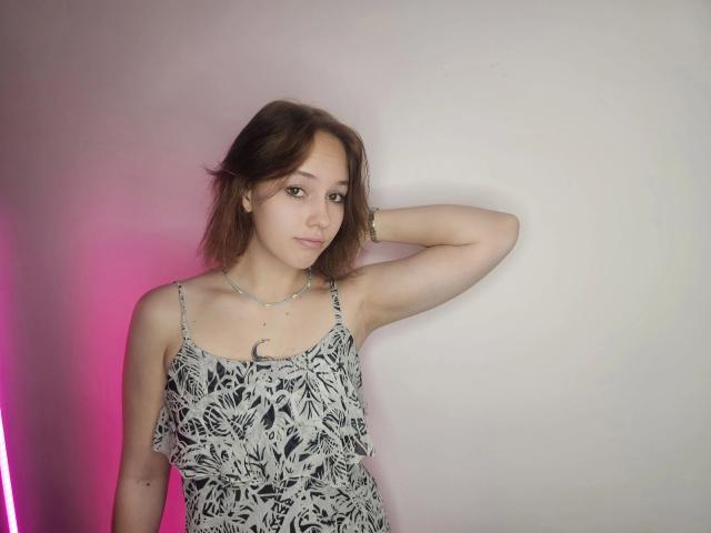 RikkiTikki-hot with short brown hair, wearing a black and white patterned dress, standing against a wall, arm raised, looking at camera, with a pink neon light.