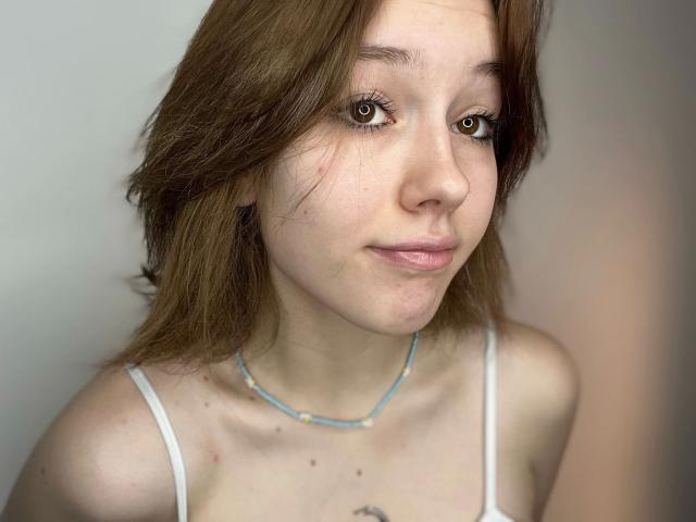 Close-up of RikkiTikki-hot with light brown, slightly messy hair. She has brown eyes with long eyelashes, a small nose, and pinkish lips. She's wearing a white spaghetti strap top and a thin blue necklace. Her skin is fair with small freckles and a visible tattoo on her chest. The image is sharp with soft, natural lighting. Her expression is neutral, slightly smiling. The background is blurred and indistinct.