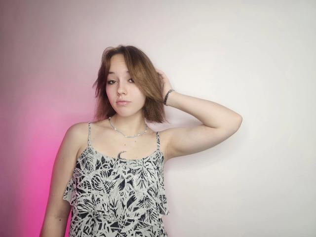 RikkiTikki-hot with shoulder-length brown hair, wearing a black and white floral dress, has a necklace and bracelet, hand in hair, pink and white light on left.