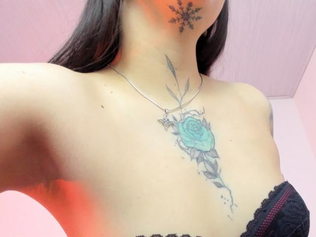 KayleeM's chest with a blue rose tattoo, black lace bra, snowflake neck tattoo, and silver cross necklace, showing her collarbone and upper chest.