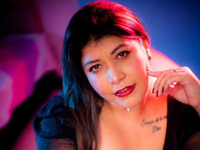 AmeliaThompson69 has red lipstick, dark hair, and a tattoo under her tits. She's looking up with a slight smile, her finger touching her chin. Red and blue lights.