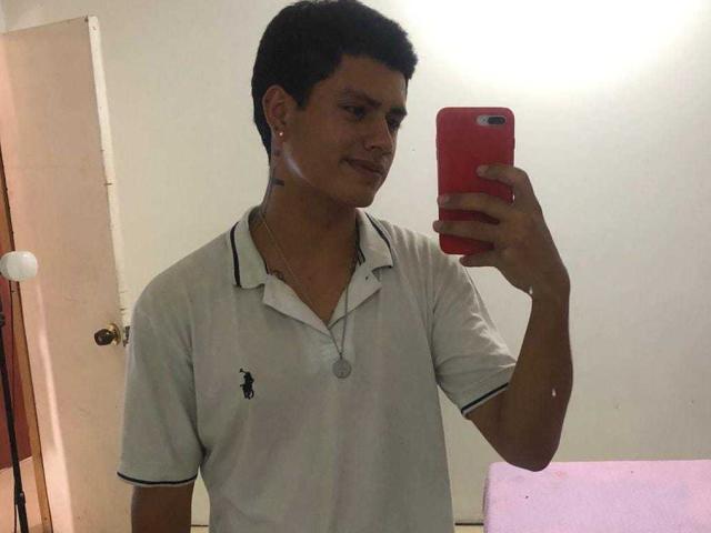 MaxRush, avec ses courts cheveux noirs, portant une chemise polo blanche, tenant un téléphone rouge, prenant un selfie devant un miroir, souriant légèrement, se tenant dans une pièce avec un sol rose..