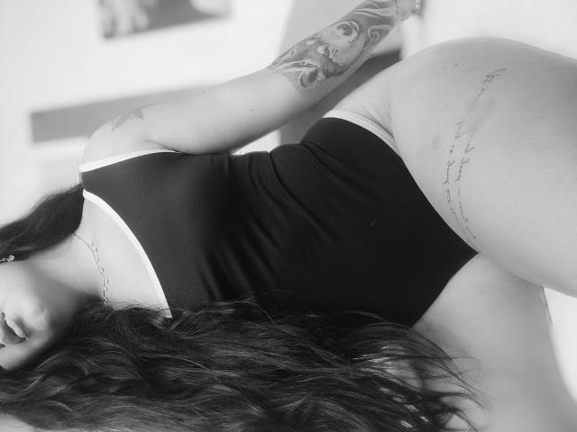 AiuniHot in a black one-piece, bent over, showing her ass and tattooed arm with long dark hair, wearing a chain necklace, in a black-and-white photo.