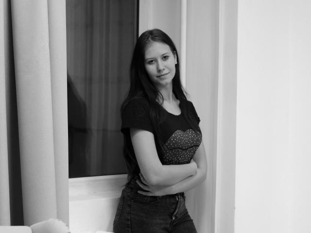 AngelicaMills with long dark hair, black shirt with sparkly chest, and jeans, arms crossed, standing by a window, smiling slightly, black and white photo.