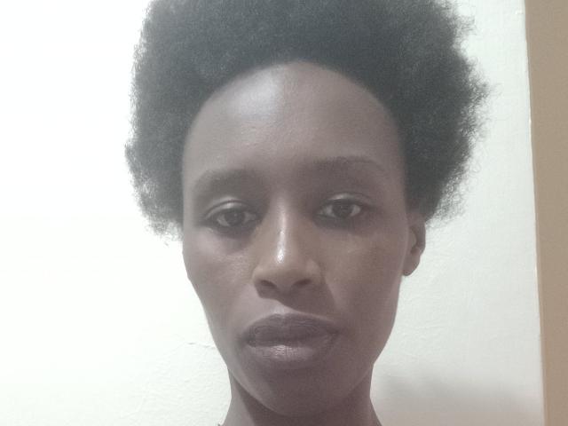 Maycee has a dark brown, afro-textured head of hair, medium brown skin, and full, slightly pouty lips with a neutral expression. Her eyes are dark and slightly squinted.