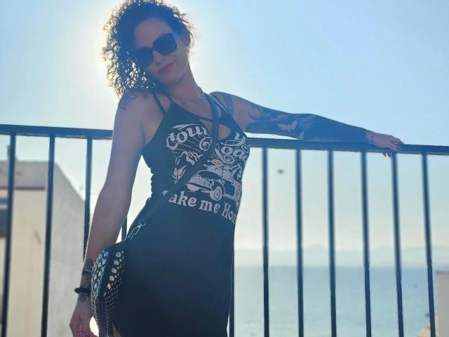 Sydney-Schmid-hot with curly hair, wearing dark sunglasses and a black dress, leaning on a metal railing, tattooed arm, sun behind, blue sky, ocean in background.