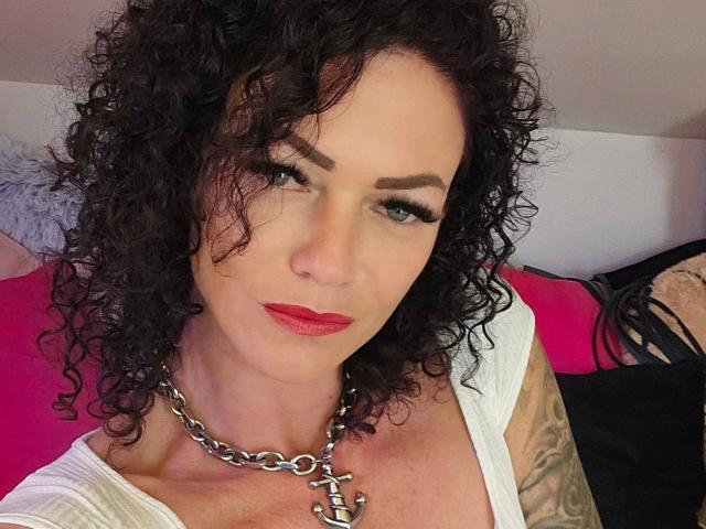 Sydney-Schmid-hot has curly black hair, red lipstick, and a chain necklace with a cross. She's wearing a white top, showing a tattoo on her right arm.