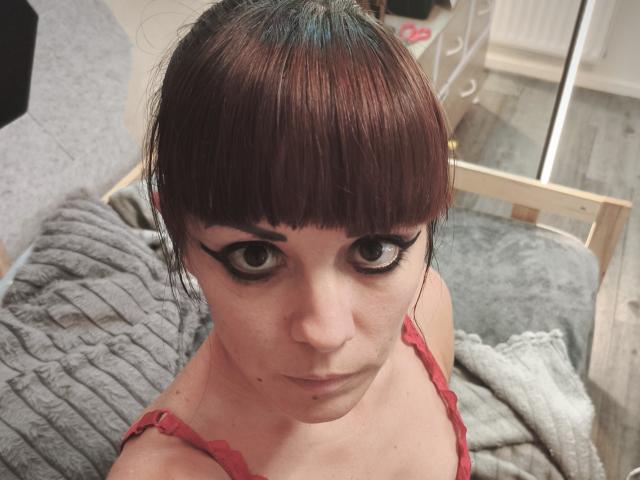 OrianaLaFrancaise with short brown hair, black eyeliner, and red bra, looking up with a serious expression, sitting on a gray bed with a wooden headboard.