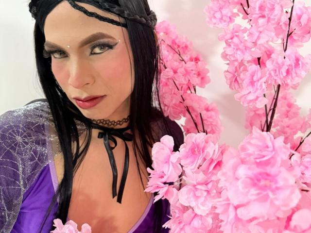 DominiqueR with black braided hair, purple dress, black choker, and pink cherry blossoms, showing off her tits and a serious, sexy look with heavy eyeliner.