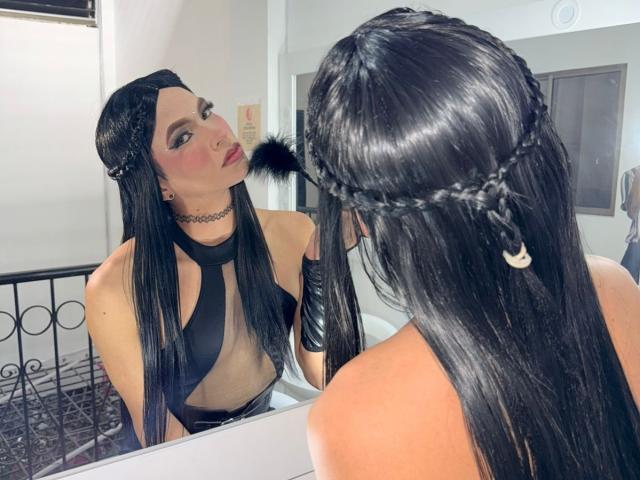 DominiqueR with long black hair in braids, wearing a black sheer top, looking at her reflection in the mirror, showing her tits and choker, in a bedroom.