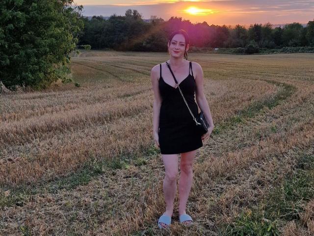 JasminWhite-hot stands in a sunlit field, wearing a black dress, white sandals, and a black crossbody bag, with a smile, and her tits are modestly visible.