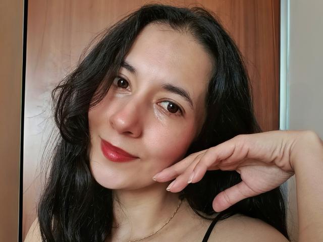 AnneStone has black wavy hair, brown eyes, red lipstick, and a slight smile, resting her hand on her cheek, with a wooden background and a gold necklace.