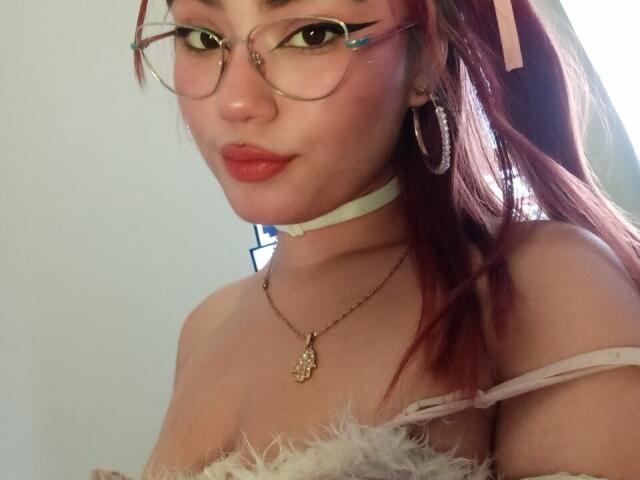 LiahMillerr with red hair, glasses, hoop earrings, white choker, necklace, and fluffy strap, pouting with red lips. Boobs visible, strap off shoulder. Soft, natural light.