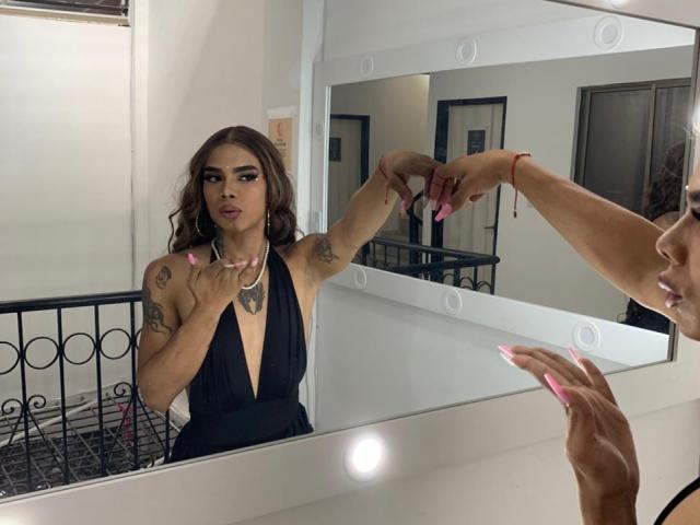 AlejaDox mit rosafarbenen Nägeln, einem schwarzen Kleid mit tiefem Ausschnitt, Tattoos und einem roten Armband, die ihre Brust berührt und sich im Spiegel betrachtet, wobei ihr Gesicht und ihre Hände reflektiert werden..