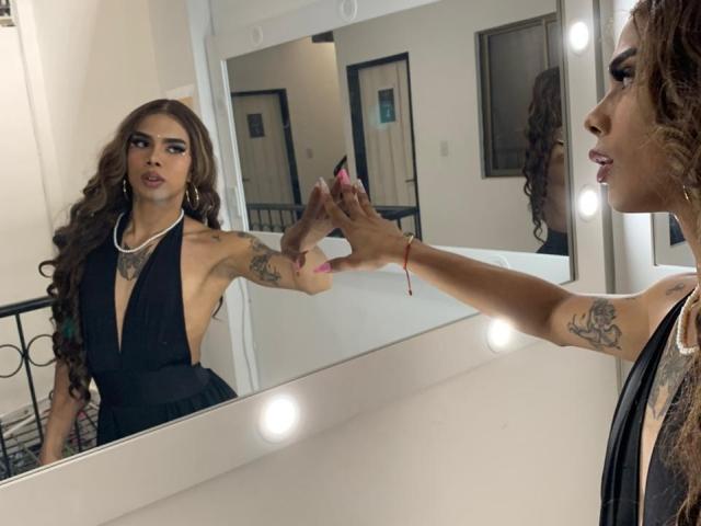 AlejaDox with long brown hair, wearing a black halter dress, tattoos on arms, looking in mirror, touching face, mirror lights, white walls, black railing, pink nail polish.