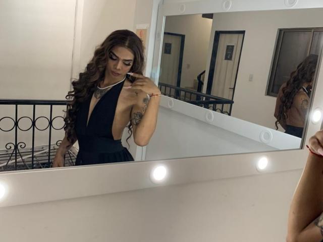 AlejaDox mit langen, braunen, gewellten Haaren, trägt ein schwarzes Top mit Halter und spiegelt sich in einem Spiegel mit Lichtern wider. Sie berührt ihr Gesicht und zeigt dabei ein Tattoo an ihrem rechten Arm..
