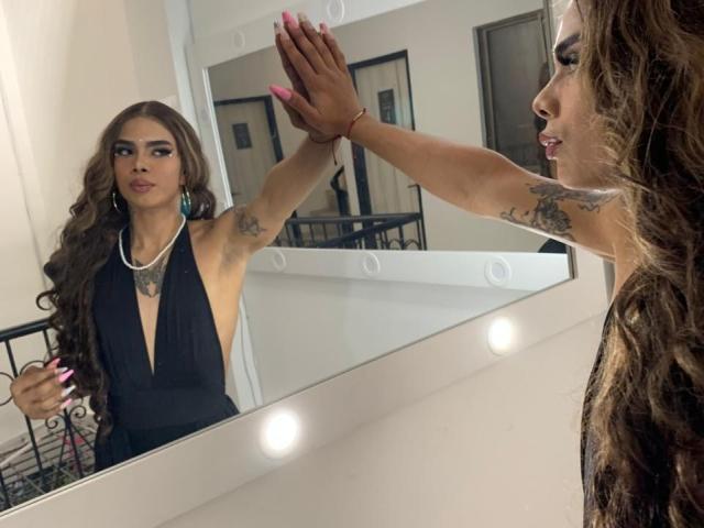 AlejaDox, avec de longs cheveux bruns bouclés, une robe noire à décolleté en V plongeant, des tatouages sur les bras, des ongles roses, un reflet dans un miroir, une main contre le miroir, un mur blanc, des lumières rondes autour du miroir, une expression sérieuse..