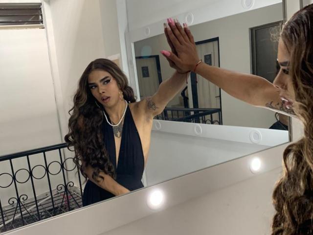 AlejaDox with long brown curly hair, wearing a black halter dress, touches her reflection's arm, showing tattoos and pink nails, in front of a mirror with bulbs.