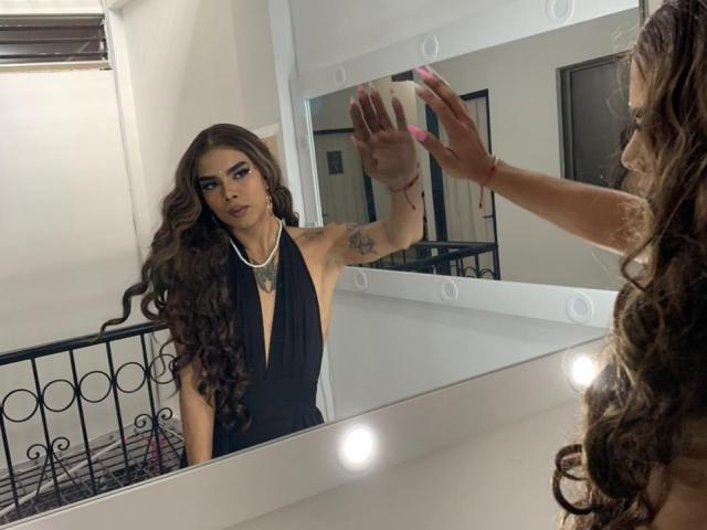 AlejaDox with long brown curls, wearing a black halter dress, stands in front of a mirror, touching it with pink-nailed hands, showing a tattoo on her right arm.