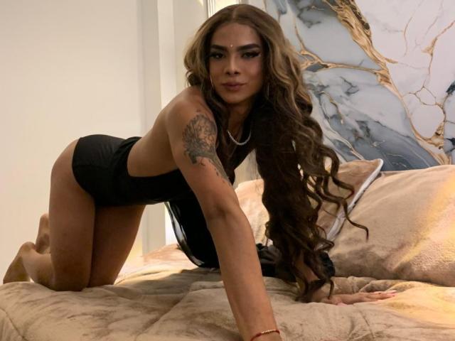 AlejaDox, avec ses longs cheveux bruns ondulés, sa lingerie noire et son tatouage sur le bras, est à quatre pattes sur un lit, regardant la caméra et montrant ses fesses..