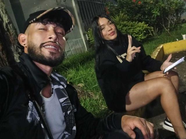 SachaAndFercho with a beard and cap, grinning, and Fercho with long black hair, pouting, wearing a black hoodie and shorts, sitting outdoors, making a peace sign.