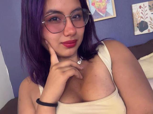 NovaAllen con cabello morado, gafas, lápiz labial rojo, camiseta blanca, tocándose la barbilla, mostrando los senos, pulsera negra, sonrisa sutil, pared azul, obra de arte enmarcada en el fondo, plano medio..