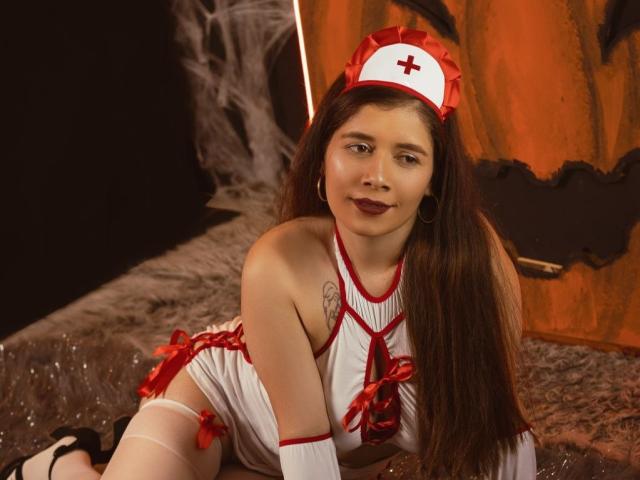 EmilyCambell with long brown hair, wearing a red and white nurse outfit, dark lipstick, and a nurse cap, kneeling, showing her tits, and a small tattoo on her right shoulder.