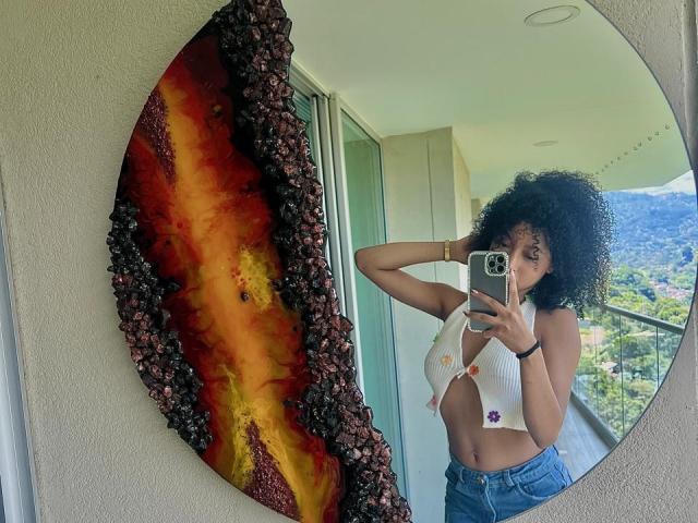 Curvy IvyRoberts with fluffy black hair, small white top showing her ass, holding a phone, standing in front of a mirror with a colorful, textured art piece.