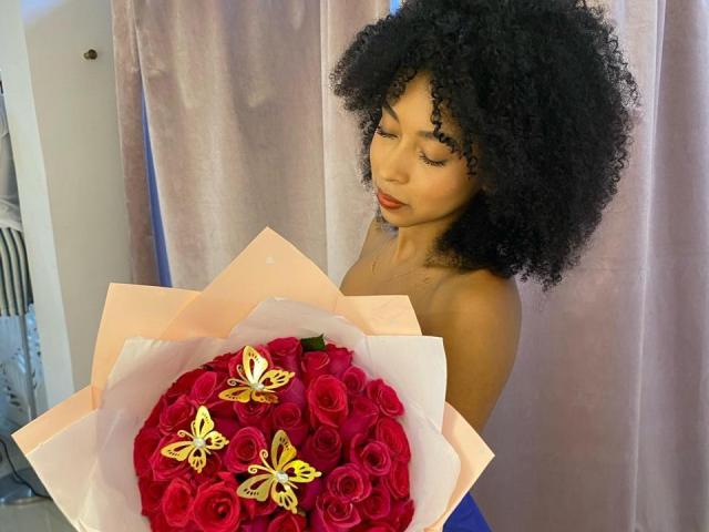 IvyRoberts with curly black hair, holding a bouquet of red roses with yellow butterflies. She's topless, showing her bare shoulders and a small tattoo on her neck.