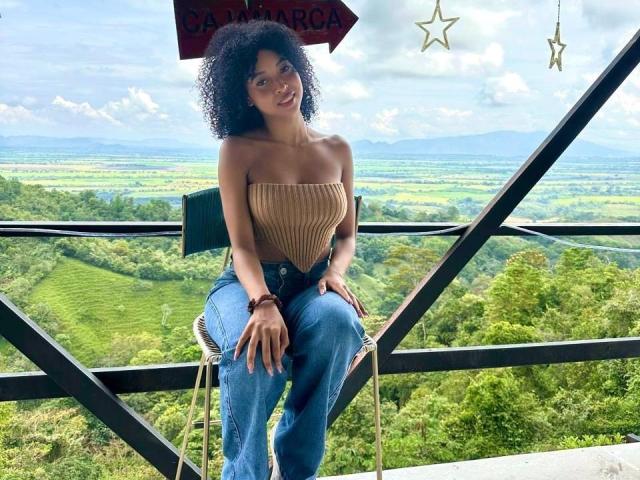 IvyRoberts with curly black hair, tan strapless top, and blue jeans, sitting on a chair, looking happy, outdoors, green landscape, mountains in the distance, star decorations above.