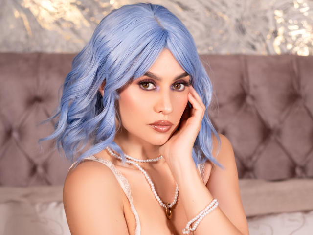 Lilaa, uma mulher de cabelos azuis, olhos castanhos e maquiagem, vestindo uma blusa de renda, um colar de pérolas e uma pulseira, tocando seu rosto, mostrando seus seios, em uma pose suave e sensual..