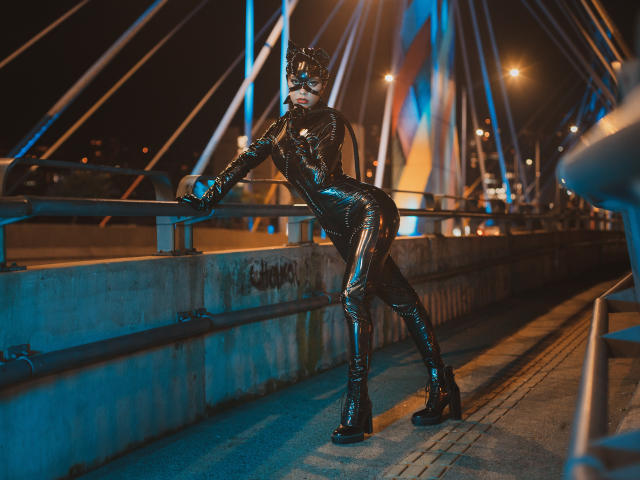 HannaSotoo in shiny black latex outfit with cat ears, standing on bridge, leaning on rail, blue and orange lights, high heels, butt facing camera, nighttime, urban setting.